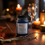 Load image into Gallery viewer, Blue hand poured candle jar labeled 'Twilight Woods' on a wooden surface with blurred candles and glasses in the background. Southern Charm Farmhouse.
