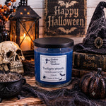 Load image into Gallery viewer, A hand-poured candle in a clear glass jar with a black lid sits on a rustic wooden surface surrounded by Halloween decor, including a skull, lantern, witch hat, spiderweb, pumpkin, and a sign that reads 'Happy Halloween.' The label shows the brand Southern Charm Farmhouse and the scent Twilight Woods.
