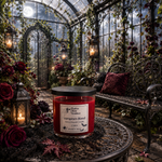 Load image into Gallery viewer, A red scented candle in a clear glass jar with a black lid sits on a round table in a gothic-style greenhouse. The label reads 'Vampire's Blood'. Southern Charm Farmhouse.
