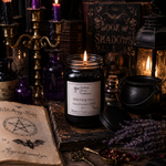 Load image into Gallery viewer, A lit candle in a 16 oz Mason  jar labeled ' Witching Hour' sits on a rustic wooden surface surrounded by occult-themed props including an open book with a pentagram, dried lavender, purple bottles, a brass candelabra, and a cauldron. Southern Charm Farmhouse.
