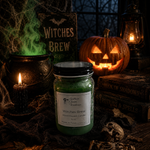 Load image into Gallery viewer, A green hand-boured candle in a glass jar labeled 'Witches Brew' by Southern Charm Farmhouse., styled on a dark table with a carved pumpkin, lantern, books, and a cauldron in a Halloween-themed setting.
