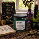 Load image into Gallery viewer, A jar candle labeled 'Witches Brew' by Southern Charm Farmhouse sits on a rustic table surrounded by witch-themed props, including a black sign reading 'Home of the Wicked Witch,' dried flowers in a purple vase, a black candle, an open book, and a spoon with herbs.
