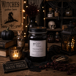 Load image into Gallery viewer, A 16 oz Mason jar hand poured candle labeled 'Witching Hour' by Southern Charm Farmhouse, styled on a rustic wooden table with Halloween decor including a small pumpkin, books, a lit candle, a lantern, skull, and dried herbs.
