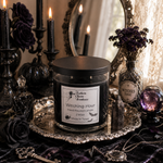 Load image into Gallery viewer, A gothic-themed hand-boured candle in a clear glass jar with a black lid, labeled 'Witching Hour' by Southern Charm Farmhouse, displayed on an ornate silver tray. The candle is surrounded by dark roses, black candles, a mirror, and a bottle of witch's tonic in a lace-draped setting.
