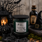 Load image into Gallery viewer, A dark green hand-poured candle in a clear glass jar with a black lid, labeled 'Witches Brew' by Southern Charm Farmhouse, styled on a rustic wooden surface with Halloween props including a lantern, black pumpkin, witch hat, potion bottle, and book.
