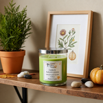 Load image into Gallery viewer, Green hand poured candle with a Cedar &amp; Autumn Leaves label on a wooden surface next to a plant, framed picture, and pumpkins.  Southern Charm Farmhouse.

