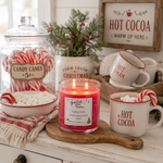 Load image into Gallery viewer, A lit red candle labeled 'Candy Cane' sits on a wooden board on a rustic white table, surrounded by holiday-themed props including a jar of candy canes, a bowl of marshmallows, a mug with marshmallows and a candy cane, and a sign that reads 'HOT COCOA'. Southern Charm Farmhouse.
