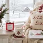 Load image into Gallery viewer, A cozy holiday living room scene with a lit red hand poured Candy Cane candle in a glass jar on a side table next to a mug of hot cocoa topped with marshmallows and candy canes. A cream knit throw blanket and embroidered pillow reading 'Let it Snow' are draped over an armchair, with a window showing a snowy outdoor view in the background. Southern Charm Farmhouse.
