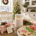 Load image into Gallery viewer, A hand-poured candle labeled 'Christmas Cookie' by Southern Charm Farmhouse is displayed on a rustic wooden table in a cozy, decorated holiday kitchen setting. The scene includes Christmas cookies on a plate, a wrapped gift with a 'Cookies, Friends, Christmas' tag, pine branches with red berries, and a decorated Christmas tree in the background.
