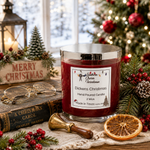 Load image into Gallery viewer, A red scented candle in a clear glass jar with a silver metal lid sits on a rustic table runner surrounded by Christmas decorations, including a book, lantern, dried orange slice, pinecones, and evergreen branches. The candle label reads 'Dickens Christmas' and shows the brand 'Southern Charm Farmhouse'.
