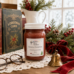 Load image into Gallery viewer, A hand-poured candle labeled Dickens Christmas in a clear glass jar with a white lid sits on a wooden table surrounded by holiday decorations, including a lace doily, a brass bell, red velvet ribbon, evergreen branches with red berries, and vintage books in the background. Southern Charm Farmhouse.
