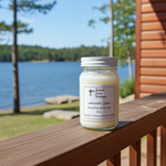 Load image into Gallery viewer, 16 oz Mason Jar of candle labeled 'Lakeside Cabin' on a wooden deck with a scenic background. Southern Charm Farmhouse.
