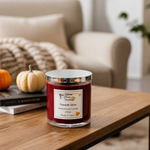 Load image into Gallery viewer, A 2-wick hand poured wax candle in a clear glass jar with a silver lid sits on a wooden table in a cozy living room setting. The candle wax is a deep red, and the label reads 'Fireside Glow' by 'Southern Charm Farmhouse'.
