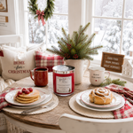 Load image into Gallery viewer, A cozy holiday table setting in a bright room, featuring a red hand poured Home 4 the Holidays candle, a red mug, a white mug reading 'Merry Christmas,' pancakes with berries, a cinnamon roll, and greenery decor near large windows. Southern Charm Farmhouse.

