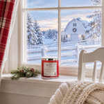Load image into Gallery viewer, A red scented hand poured Home 4 the Holidays candle in a glass jar sits on a snowy windowsill, with a white farmhouse house, snow-covered trees, and a red plaid curtain visible outside. A small pine sprig rests beside the candle, and a cream knit blanket and white chair are in the foreground. Southern Charm Farmhouse.
