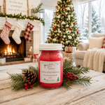 Load image into Gallery viewer, A red hand poured Home 4 the Holidays candle in a clear glass jar with a white lid sits on a rustic wooden table, surrounded by pine branches, a pinecone, and red berries. The background shows a cozy, festive living room with a lit fireplace, stockings, and a decorated Christmas tree. Southern Charm Farmhouse.
