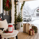 Load image into Gallery viewer, A large red Home 4 the Holidays hand poured candle in a white-labeled glass jar sits on a small white table on a snowy porch, with a cream suitcase, olive duffel bag, and wrapped gift box beside it. A wreath, garland, lantern, and snow-covered outdoor scene are visible in the background. Southern Charm Farmhouse.
