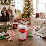 Load image into Gallery viewer, A hand poured candle in a glass jar labeled Home 4 the Holidays sitting on a wooden table in a cozy holiday living room, surrounded by wrapped gifts, a white mug that says 'Merry Everything,' and Christmas decorations in the background. Southern Charm Farmhouse.
