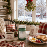 Load image into Gallery viewer, A cozy holiday breakfast scene by a window with a green hand poured candle jar labeled 'Mistletoe (type)' in the center, surrounded by a mug reading 'Good Morning Christmas,' a teapot, toast, eggs, berries, and plaid pillows. Southern Charm Farmhouse.
