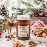 Load image into Gallery viewer, A jar of Southern Christmas Farmhouse Reindeer Munch hand poured candle sits on a snowy surface surrounded by holiday decorations, including a deer figurine, pine branches, red berries, pretzels, popcorn, and a bowl of mixed snacks. The candle jar has a white lid and a white label with the brand name and product name clearly visible.
