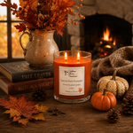 Load image into Gallery viewer, A lit spiced ram pumpkin scented hand poured candle in a clear glass jar sits on a rustic wooden table in a cozy fall setting. The scene includes autumn leaves, pinecones, small pumpkins, stacked books, a ceramic pitcher with dried leaves, and a warm fireplace in the background. Southern Charm Farmhouse.
